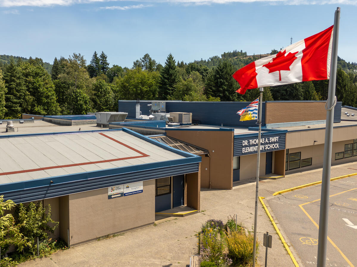Elementary school exterior with Canadian and BC flags blowing in the wind. Backdrop behind school is green trees and blue sky.
